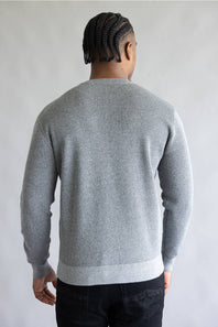 A man viewed from behind wearing a light gray ribbed knit sweater and dark pants, standing against a plain light gray background.