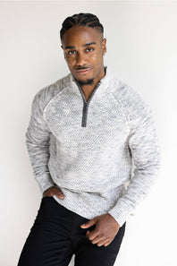 A man wearing a light gray and white marled knit quarter-zip sweater with black pants, standing against a plain white background.