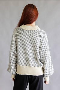 A person shown from the back wearing a cream-colored knit sweater with a black geometric lattice pattern and black pants, set against a plain gray studio background.