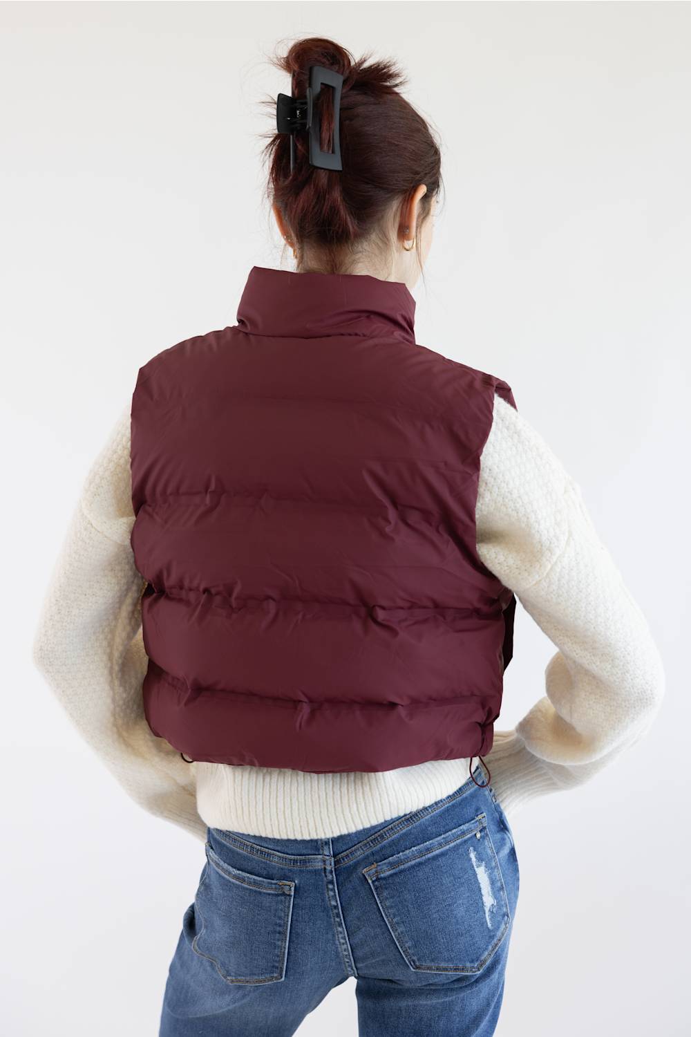 A woman shown from behind wearing a sleeveless puffer vest in burgundy over a cream knit sweater and blue jeans, against a plain white studio background.