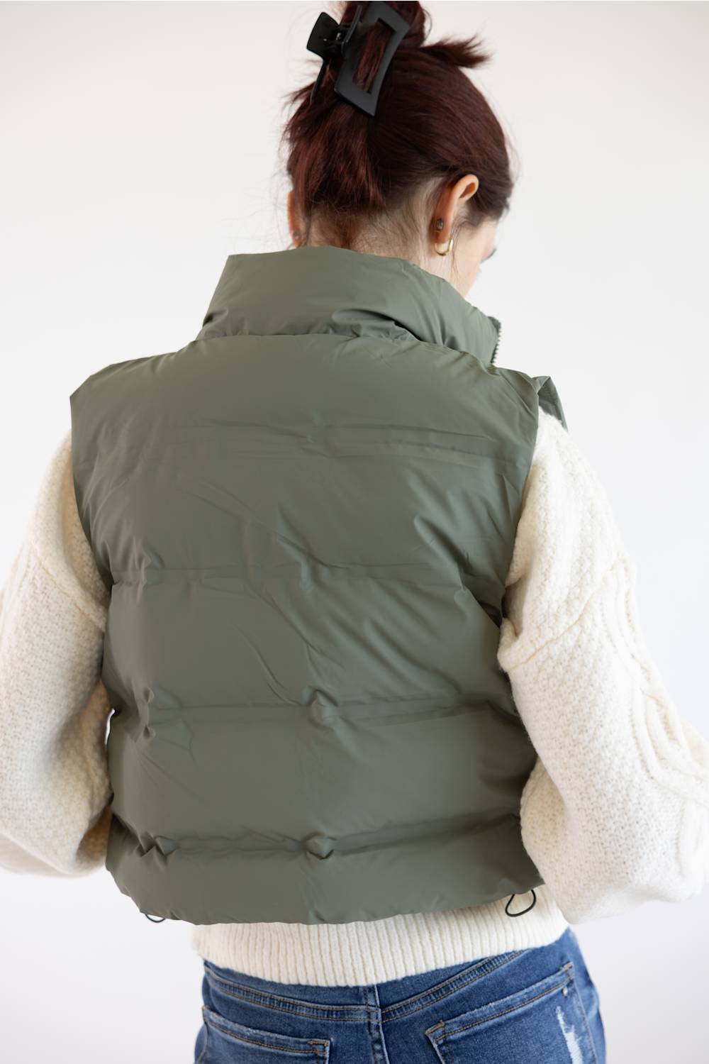 A woman viewed from behind wearing an olive green puffer vest over a cream cable-knit sweater and blue jeans, with her hair in a black claw clip against a plain white background.