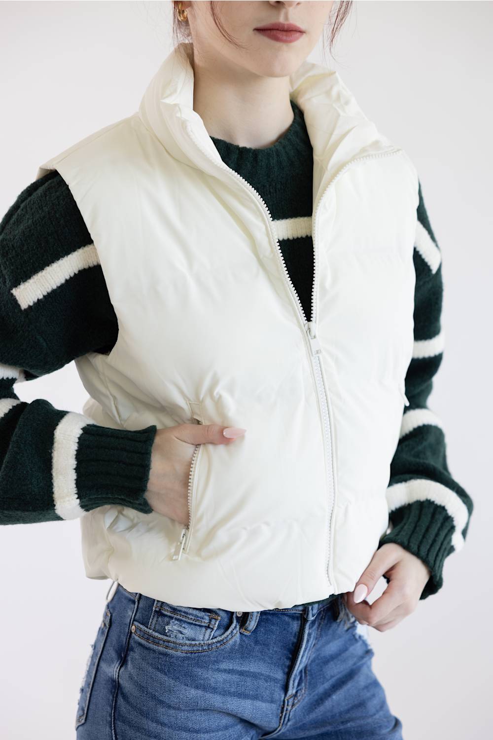 A woman wearing a cream-colored sleeveless puffer vest over a dark green and white striped knit sweater, paired with blue jeans, against a plain white background.