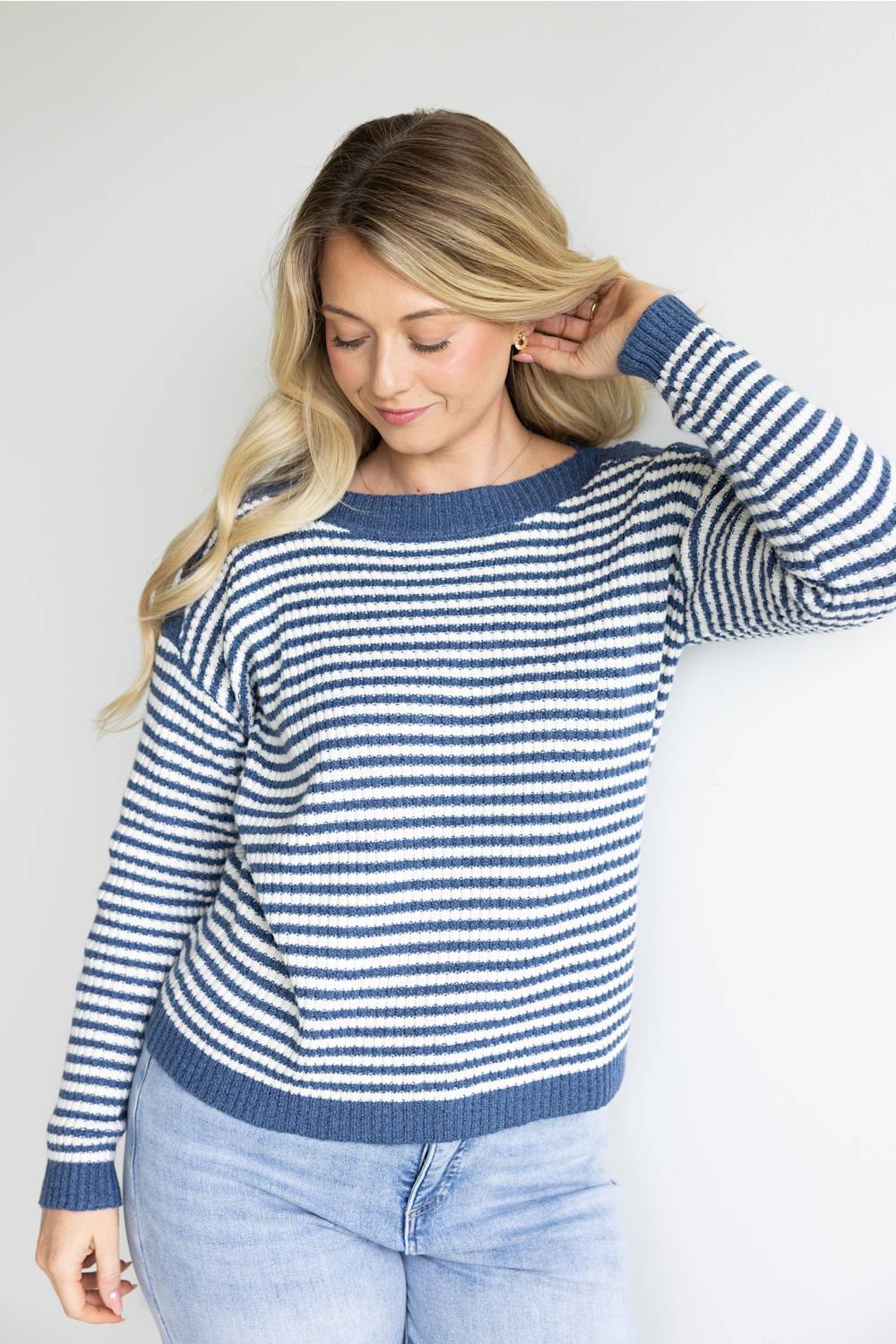 A woman wearing a blue and white horizontally striped knit sweater with a crew neck and ribbed cuffs, paired with light-wash jeans, standing against a plain light gray background.