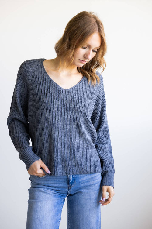 A woman wearing a slate blue ribbed knit sweater and blue jeans, standing against a plain white background.