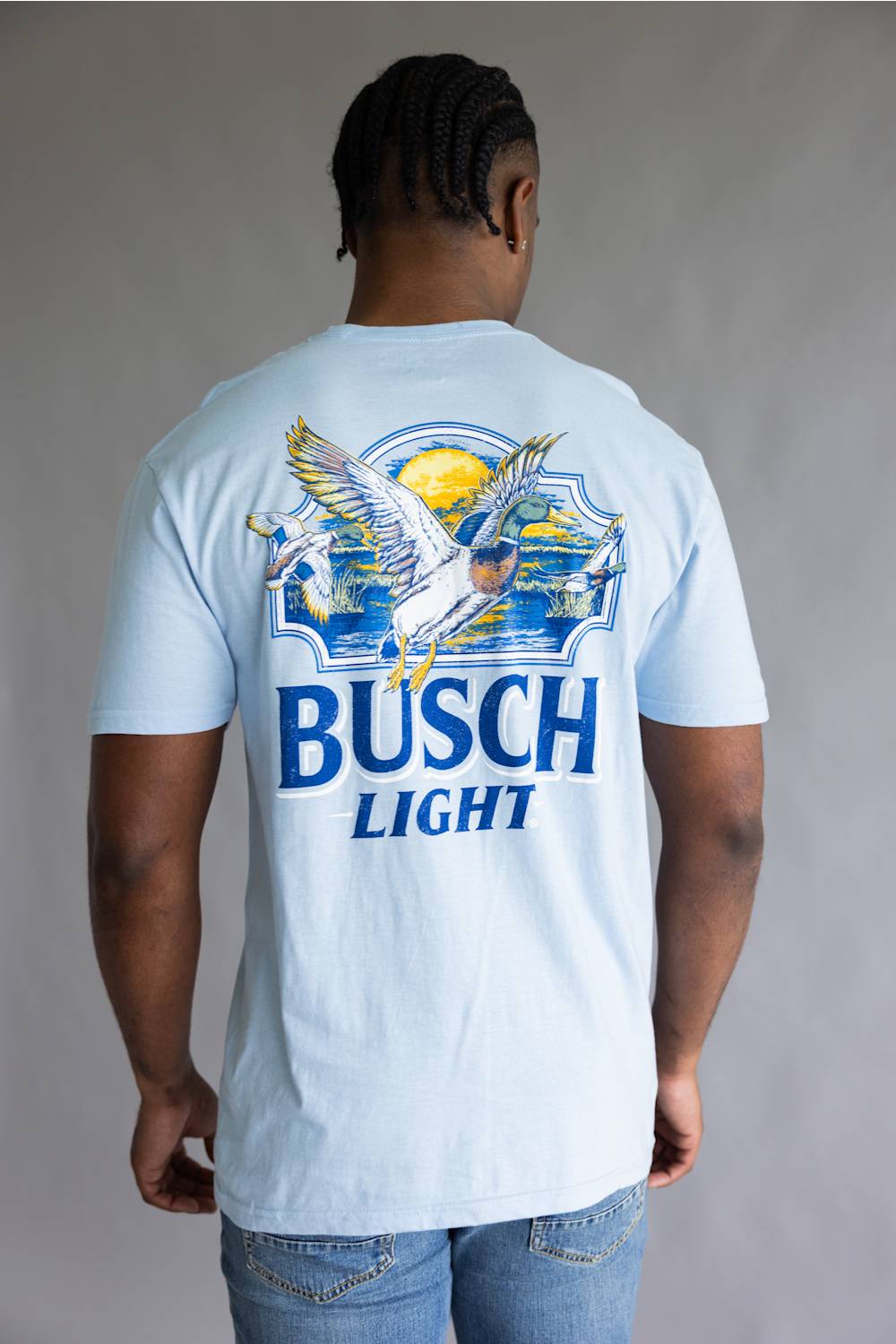 A man viewed from behind wearing a light blue crewneck t-shirt with a large graphic on the back. The graphic features the words 'BUSCH LIGHT' in blue, a white goose with yellow wings, and a yellow sun over blue waves.