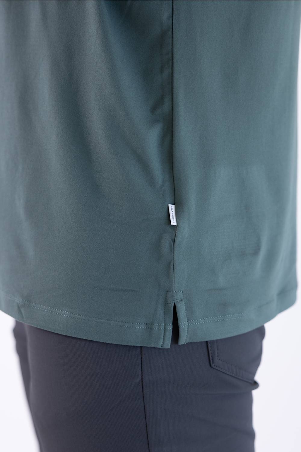 Close-up view of the back hem and side split of the 7Diamonds Core Crew T-Shirt in Drake green, showing the branded woven label tag.