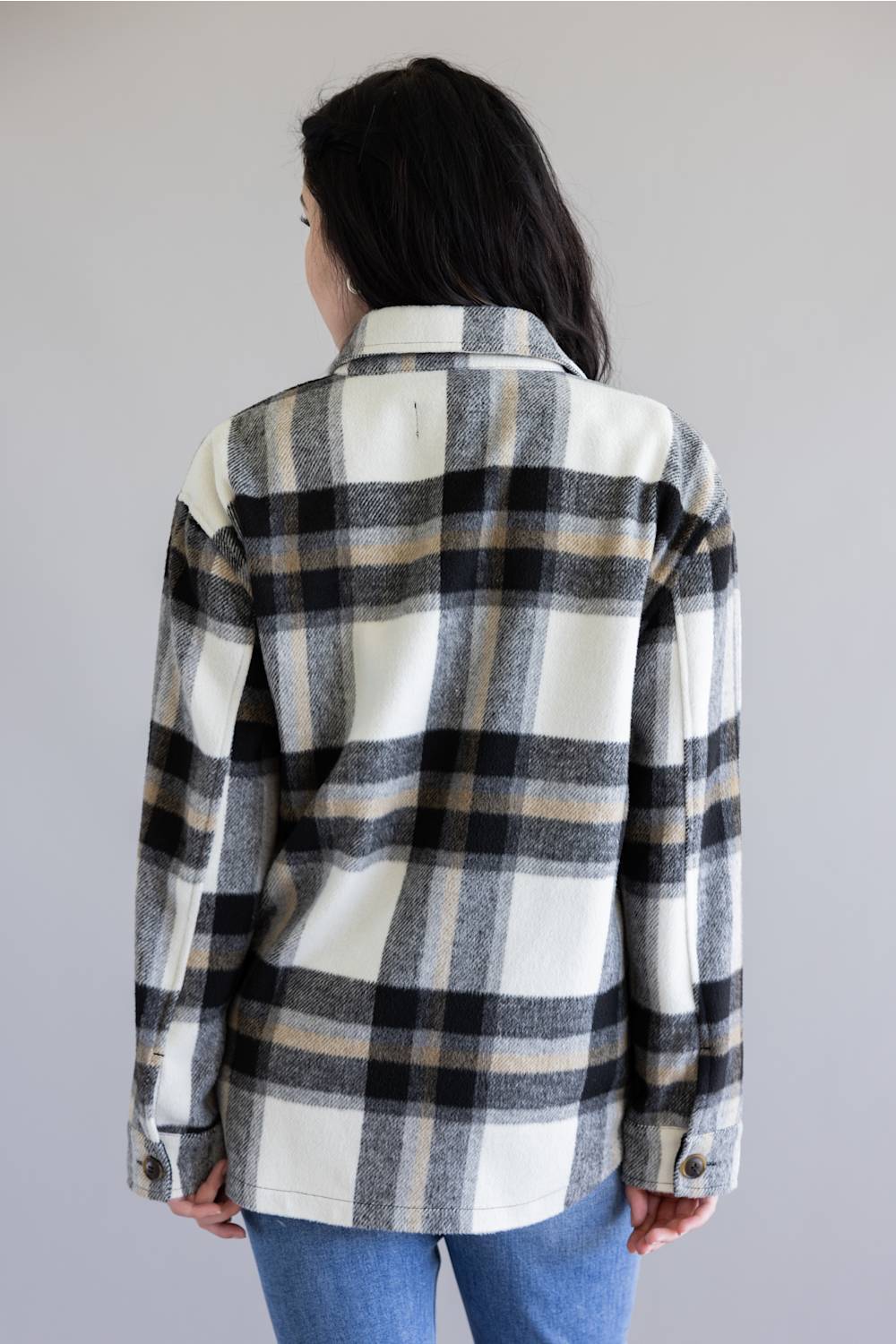 A woman viewed from behind wearing a long-sleeve plaid flannel shirt in white, black, and beige, paired with blue jeans against a plain light gray background.