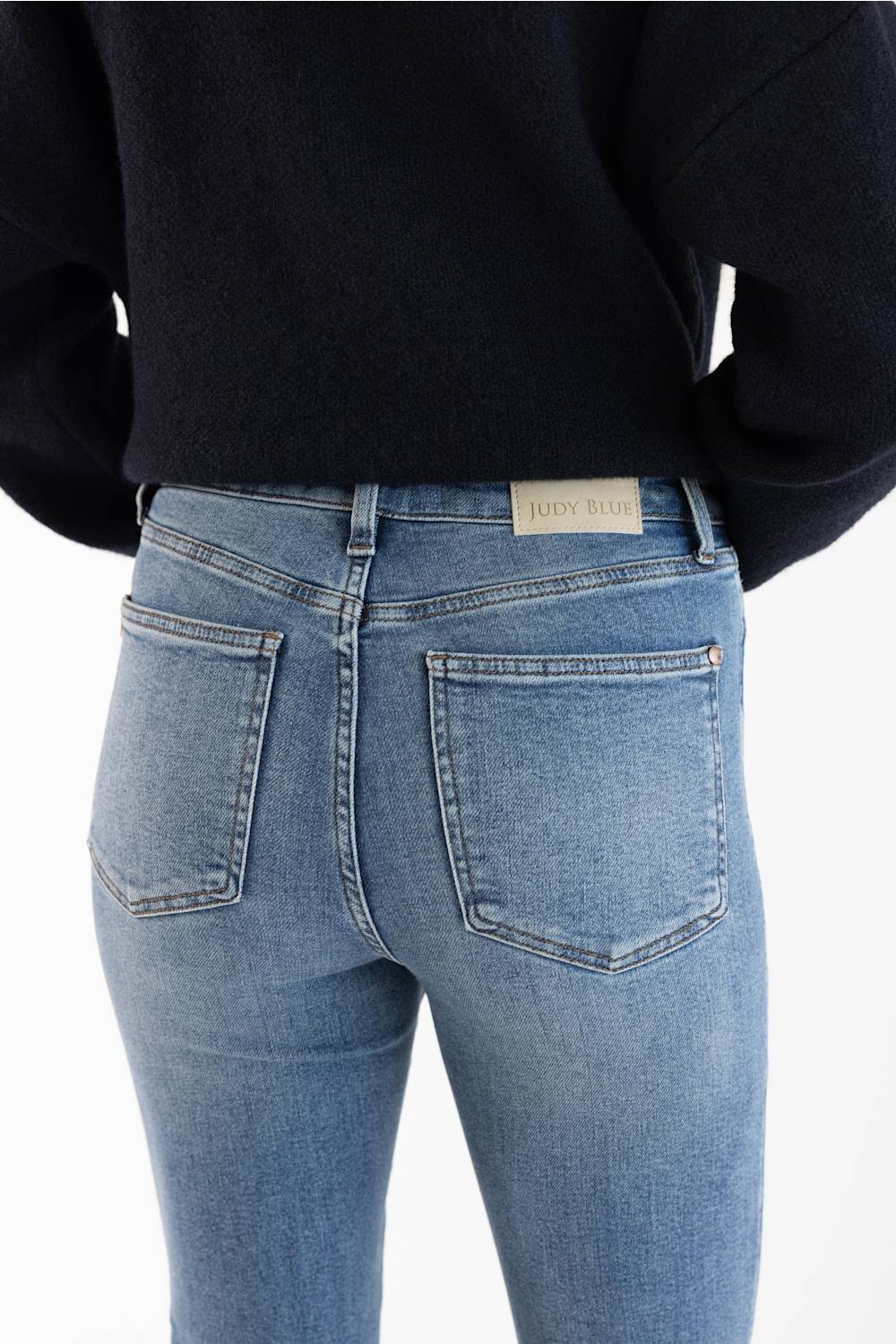 Rear view of a person wearing light blue skinny jeans and a dark navy sweater, showing the back pockets and a small 'Judy Blue' brand label on the waistband.