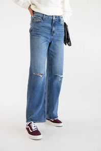 A person wearing light blue wide-leg distressed jeans with ripped knees, styled with a white ribbed sweater, burgundy sneakers, and a black clutch bag against a plain white studio background.