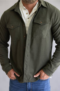 A man wearing an olive green long-sleeve button-up shirt jacket over a cream button-up shirt and blue jeans, shown from the neck down against a plain light gray background.