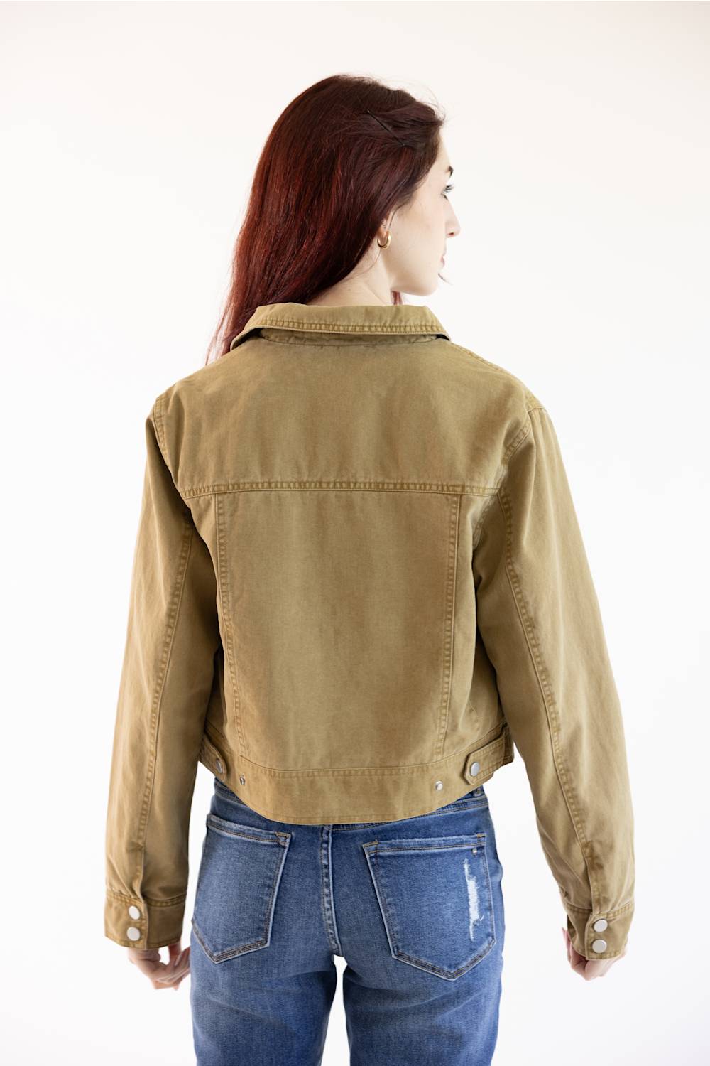 A woman with long auburn hair is shown from the back wearing a cropped tan utility jacket and blue jeans against a plain white background.