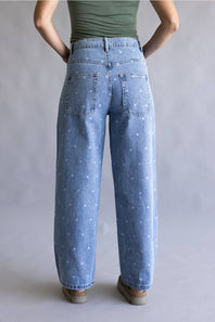 A person shown from the back wearing light blue wide-leg denim jeans with a small white star pattern, paired with a green top and brown shoes, against a neutral gray studio background.