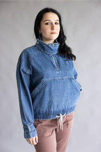 A woman modeling a blue denim half-placket pullover jacket with a mock neck and drawstring hem, paired with dusty rose pants, against a neutral gray studio background.