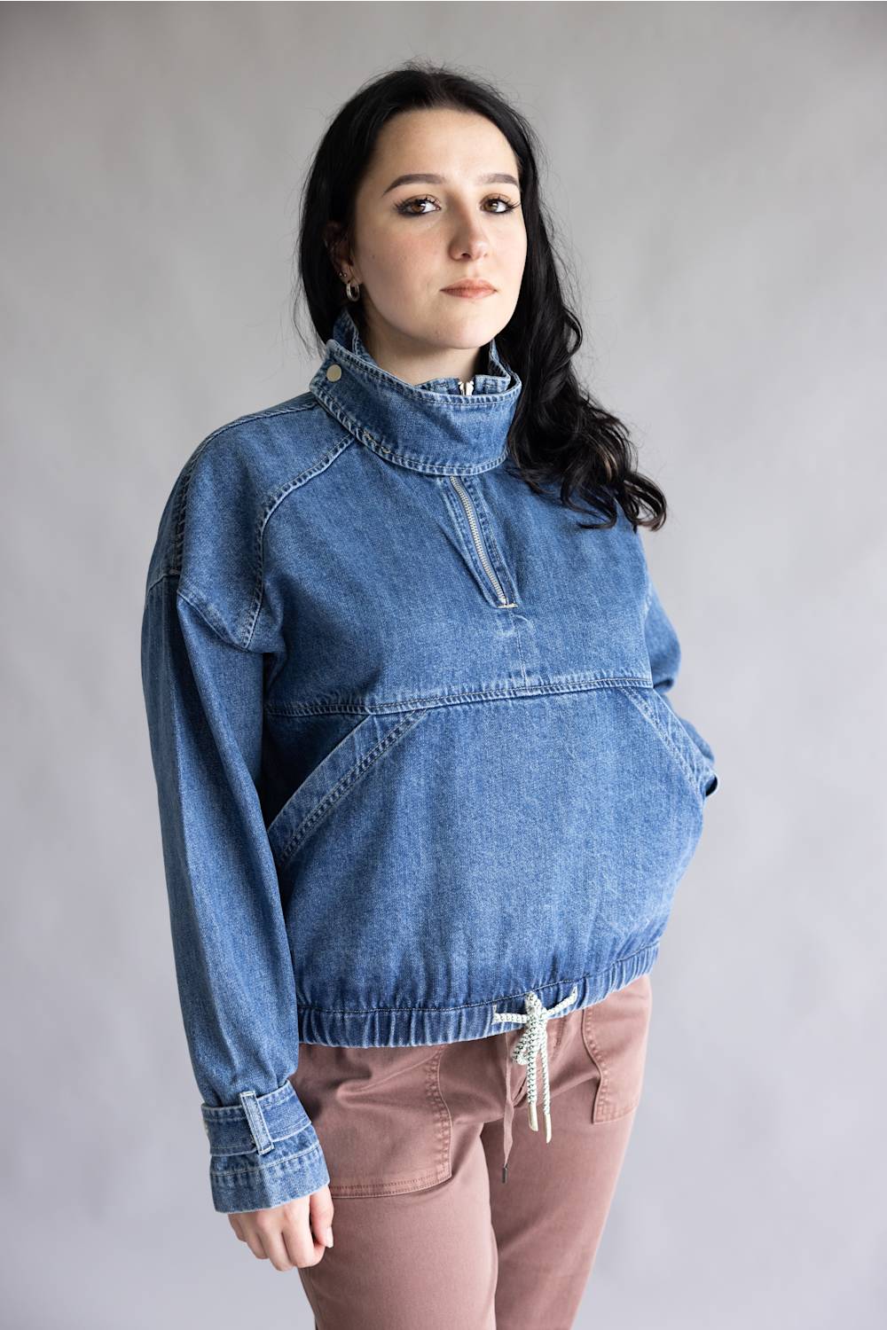 A woman modeling a blue denim half-placket pullover jacket with a mock neck and drawstring hem, paired with dusty rose pants, against a neutral gray studio background.