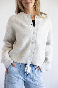 A woman modeling a light gray knit cardigan with a button-up front and drawstring detail, paired with light blue jeans against a plain white background.
