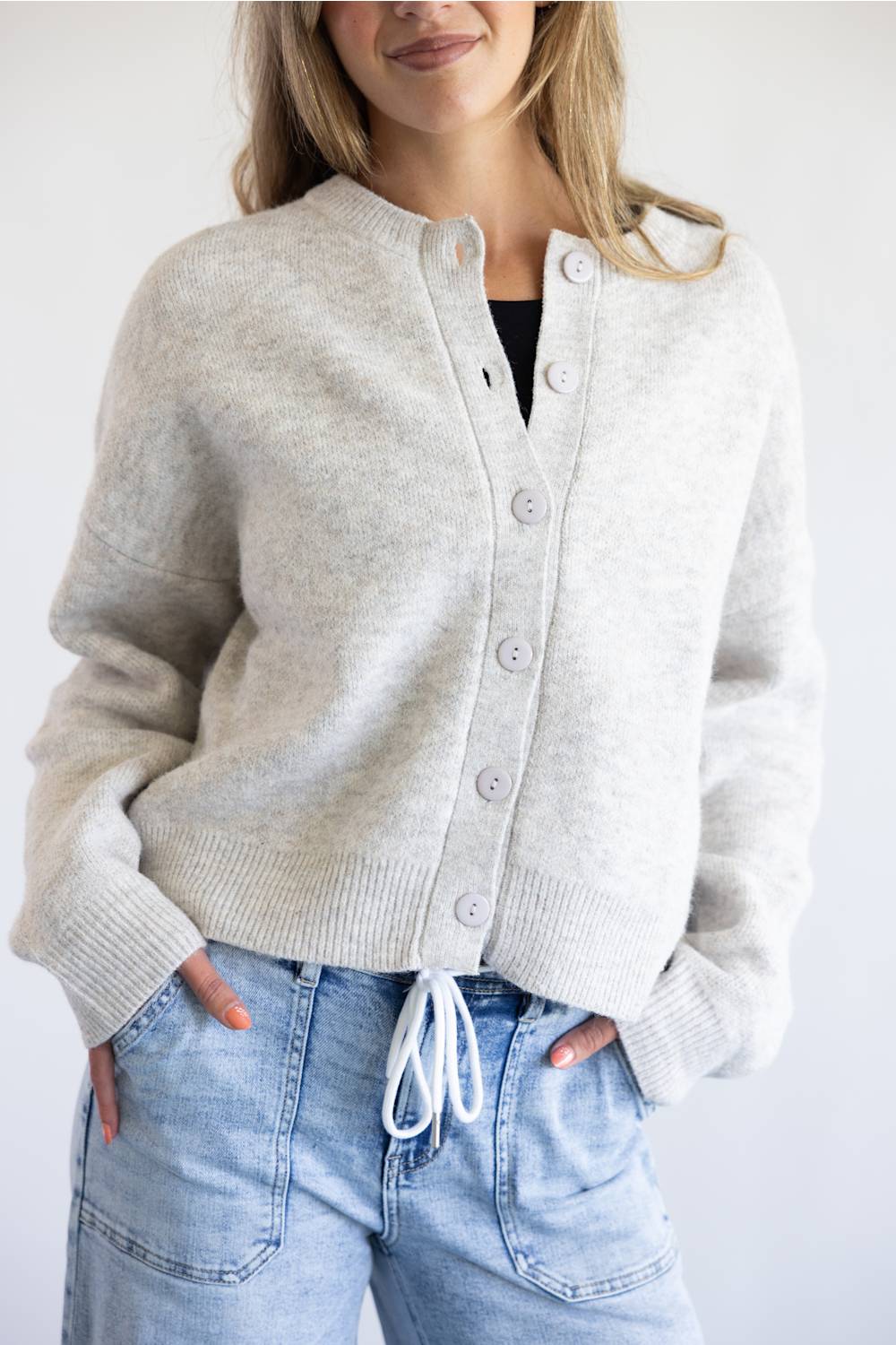 A woman modeling a light gray knit cardigan with a button-up front and drawstring detail, paired with light blue jeans against a plain white background.