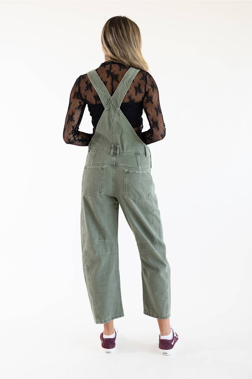 Back view of model wearing Aaron & Amber Slouchy Denim Overalls for Women in Olive featuring crossed back shoulder straps, high back patch pockets, and a slouchy barrel leg silhouette paired over a black lace long-sleeve top.