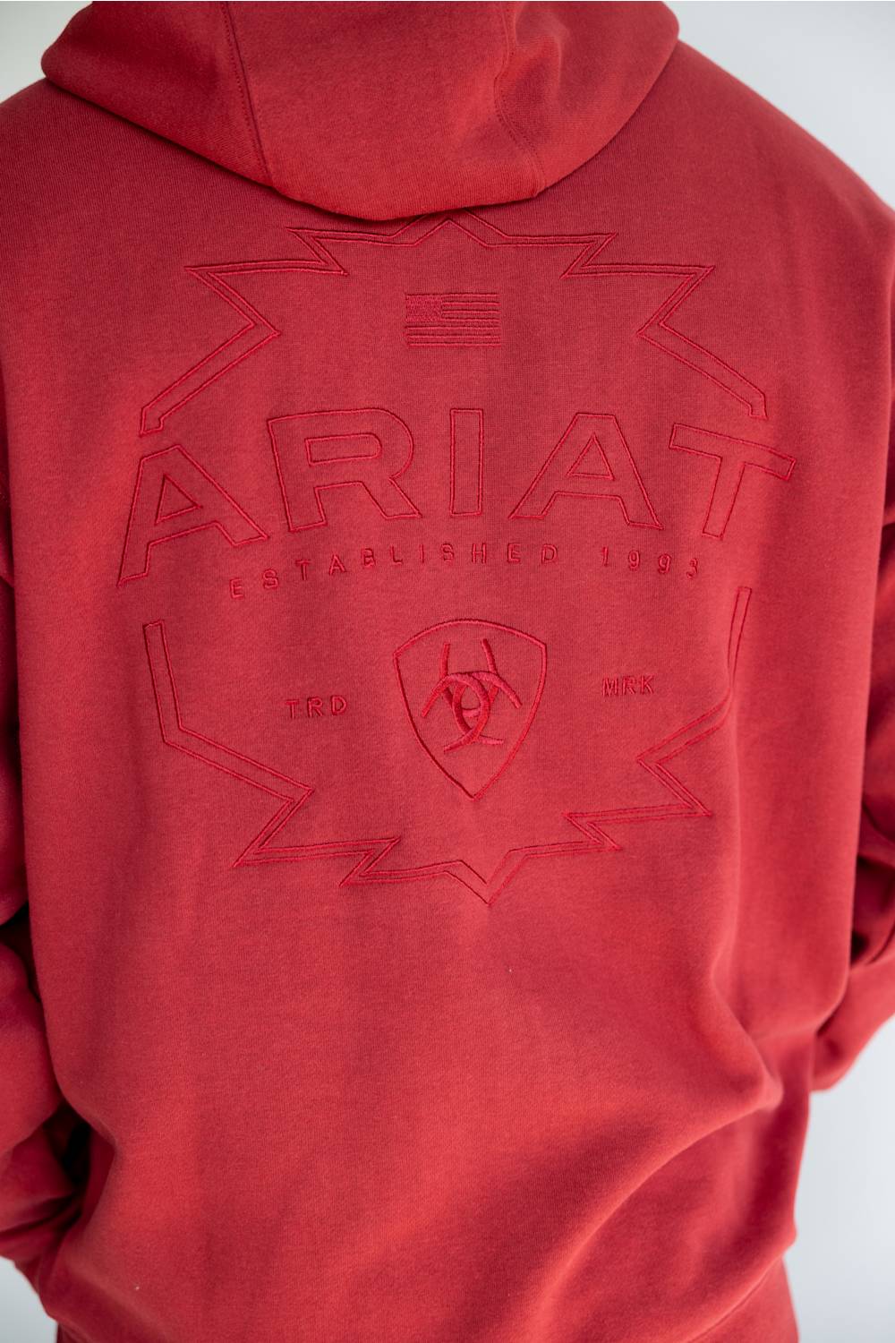 A close up of a man viewed from the back wearing a red pullover hoodie with a tonal embroidered logo across the back, photographed against a plain white studio background.