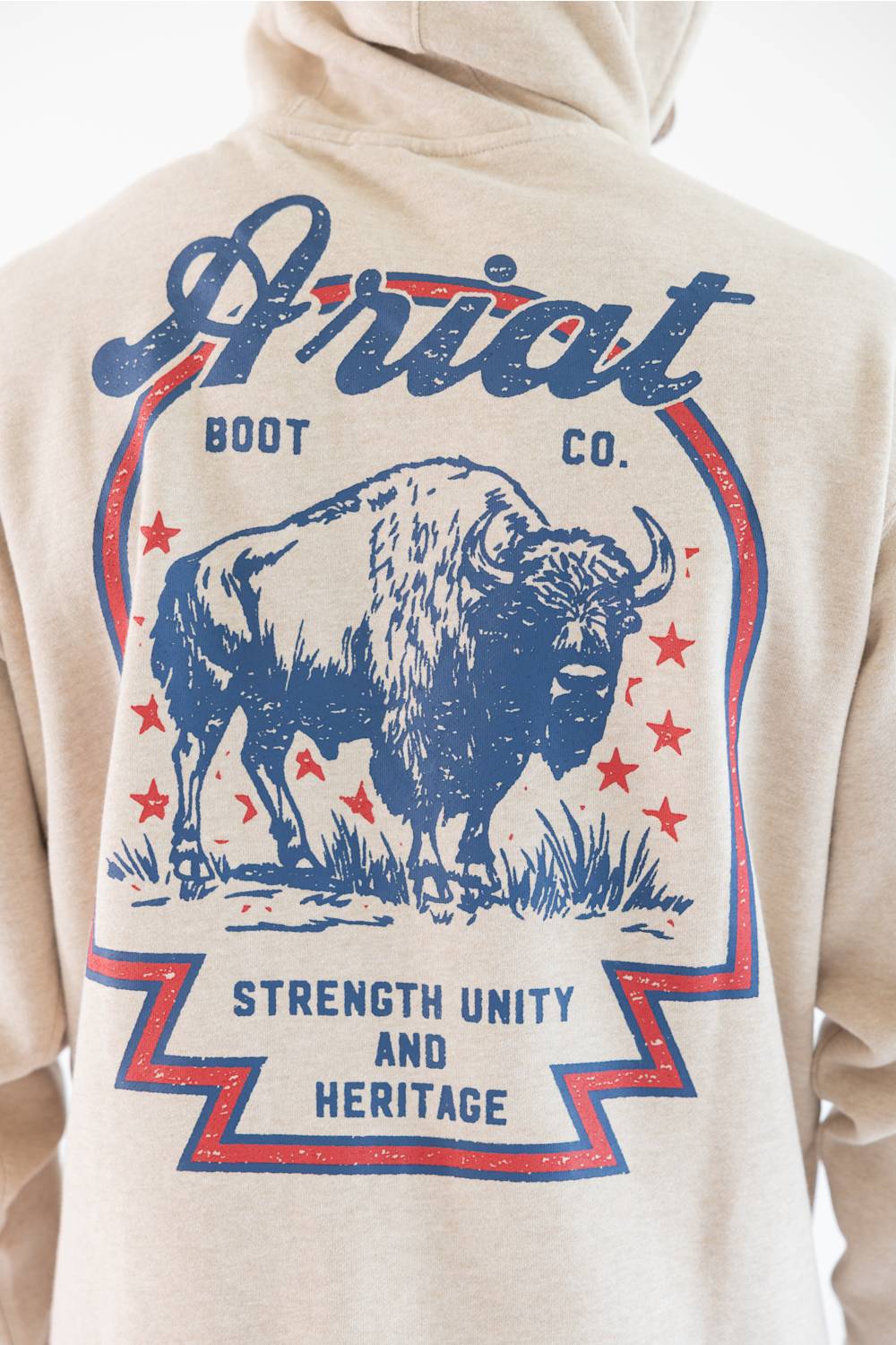 Close-up of the back of a beige hoodie featuring a large blue and red graphic print of a bison with the text 'Ariat BOOT CO.' and 'STRENGTH UNITY AND HERITAGE'.