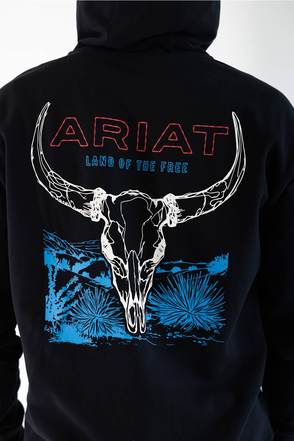 Black hoodie shown from the back, featuring a large graphic print of a white horned skull over blue desert-style artwork, with 'ARIAT' in red outlined letters and 'LAND OF THE FREE' in blue text.