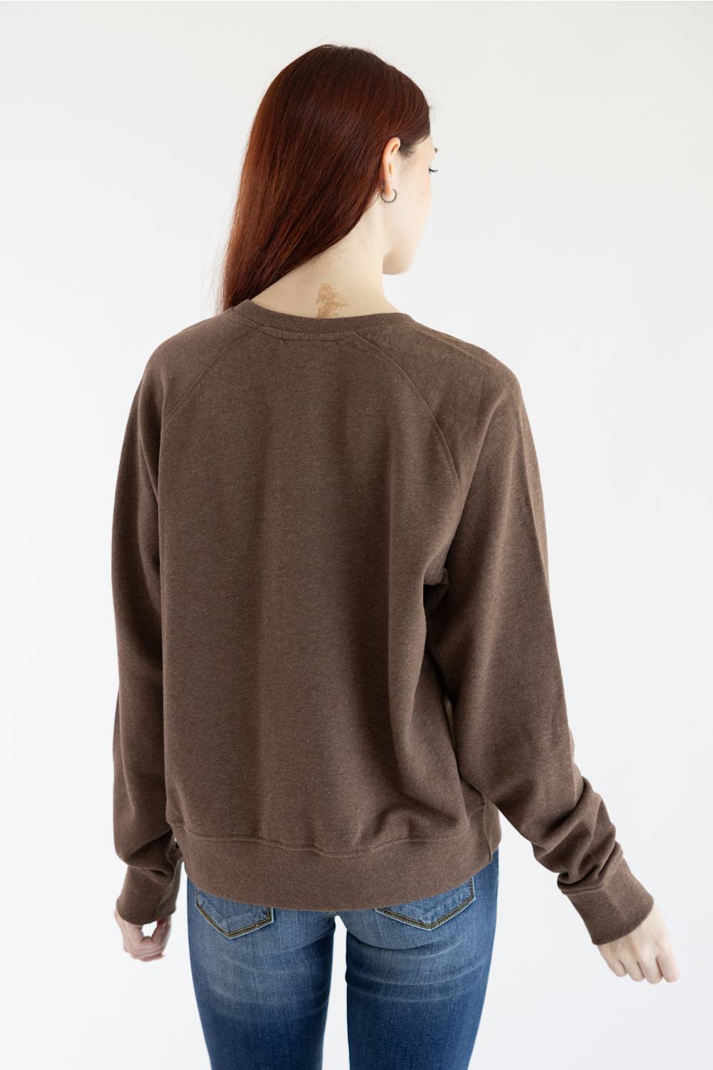 A woman with red hair wearing a brown crewneck sweatshirt with 'ARIAT' printed in yellow across the chest, paired with blue jeans, against a plain white background.