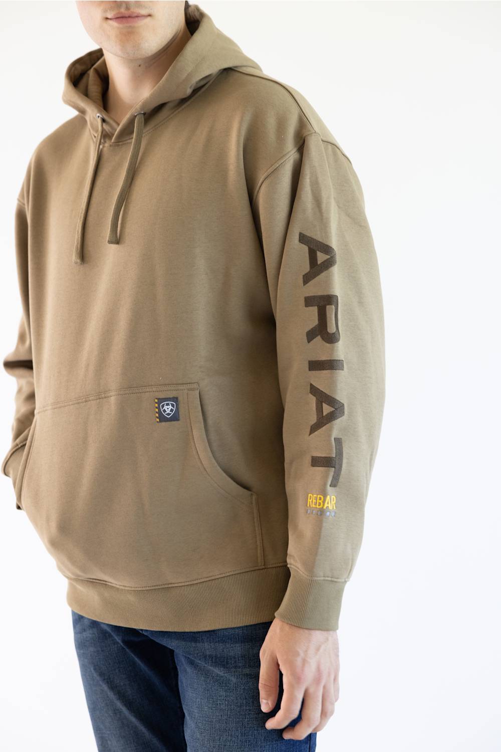 A man wearing a tan pullover hoodie with 'ARIAT' embroidered vertically in brown on the right sleeve and a small yellow logo patch near the cuff, paired with blue jeans against a plain white background.