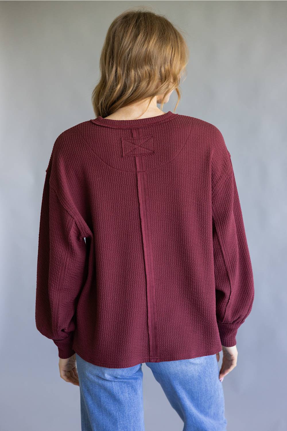 A woman viewed from behind wearing a maroon ribbed knit sweater with balloon sleeves and a central seam detail, paired with light blue jeans, against a neutral gray background.