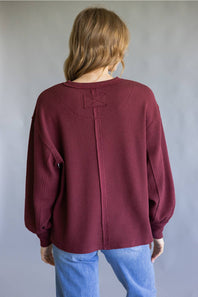A woman viewed from behind wearing a maroon ribbed knit sweater with balloon sleeves and a central seam detail, paired with light blue jeans, against a neutral gray background.