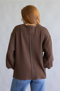 A person viewed from the back wearing a brown textured knit sweater and blue jeans against a plain light gray background.
