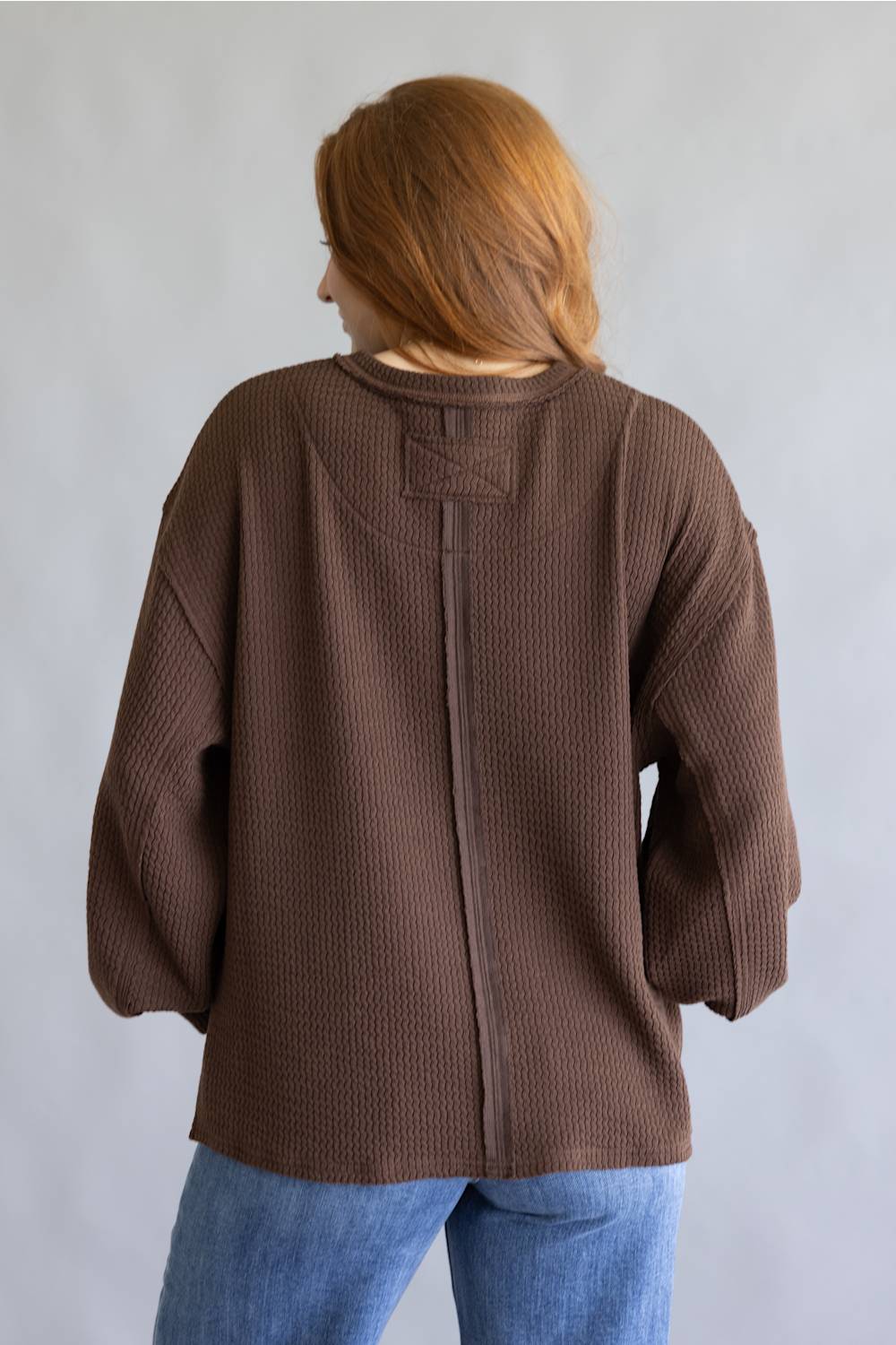 A person viewed from the back wearing a brown textured knit sweater and blue jeans against a plain light gray background.