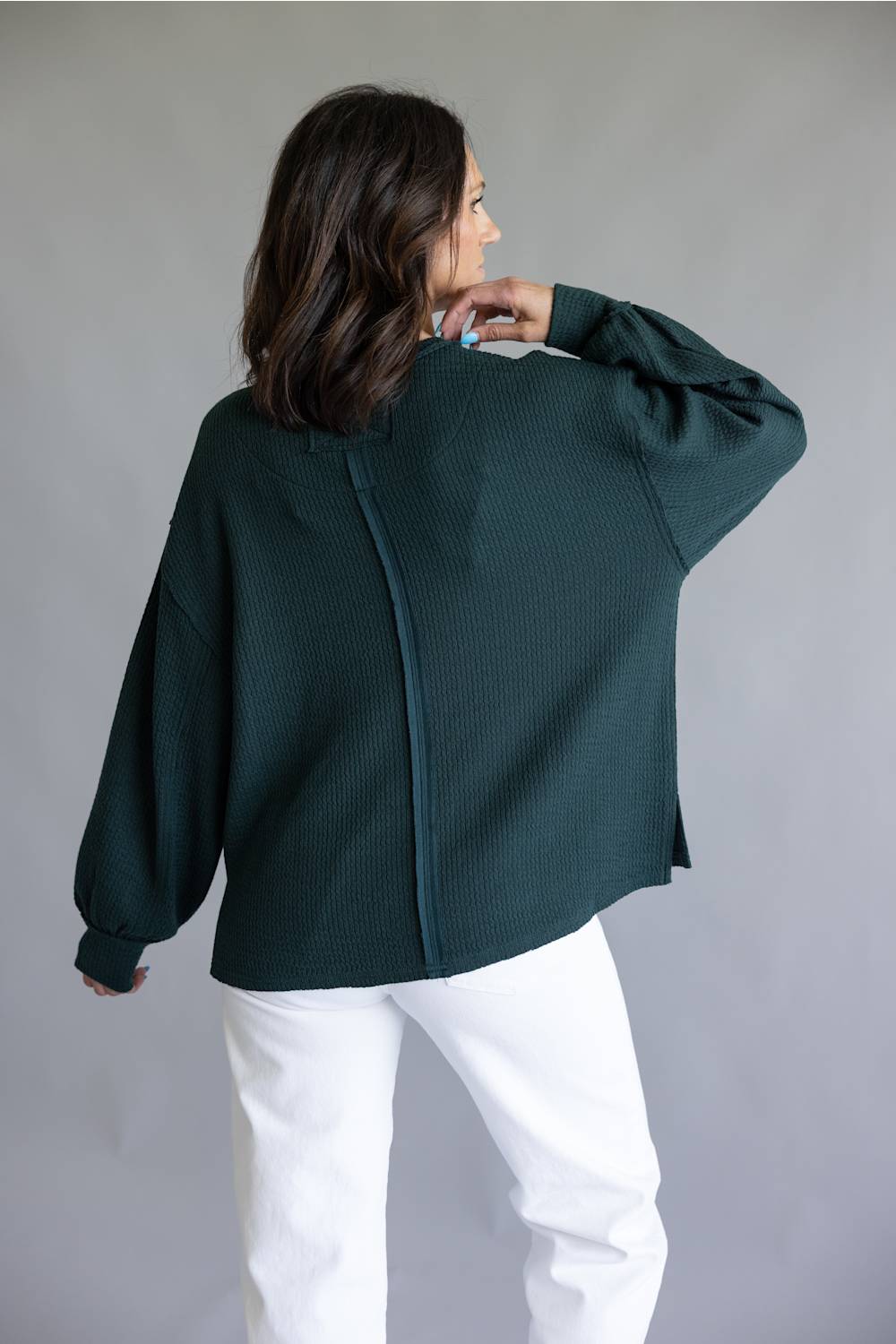 A woman wearing a dark green ribbed knit cardigan with a unique back zipper detail, paired with white pants, photographed against a neutral gray studio background.