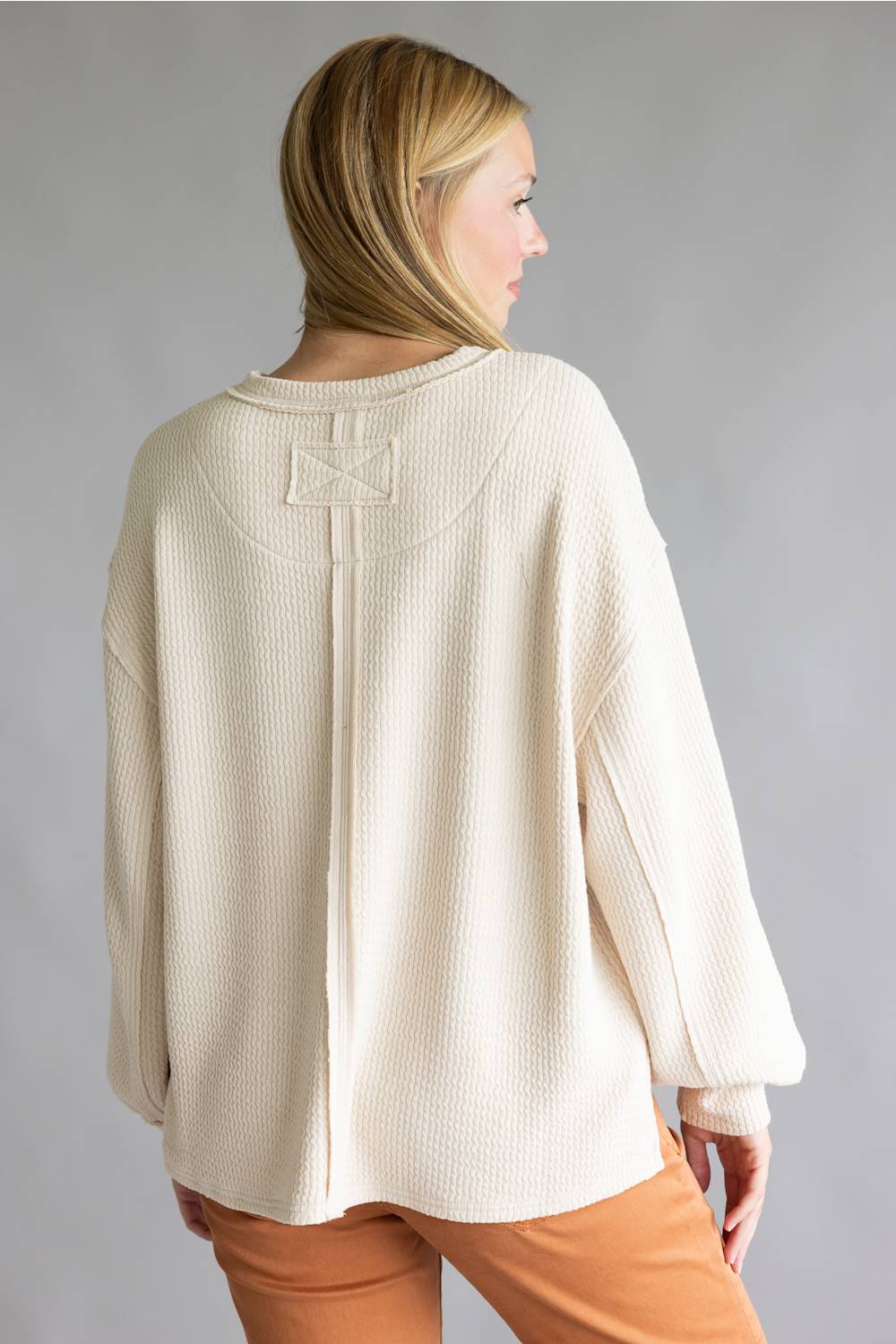 A woman shown from the back wearing a cream-colored textured knit sweater with a unique stitched detail on the upper back, paired with rust-orange pants, against a solid gray studio background.
