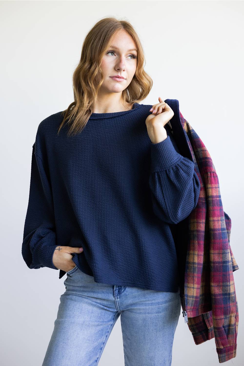 A woman wearing a navy blue waffle-knit sweater and light wash blue jeans, holding a red and blue plaid flannel shirt over her shoulder against a plain white background.