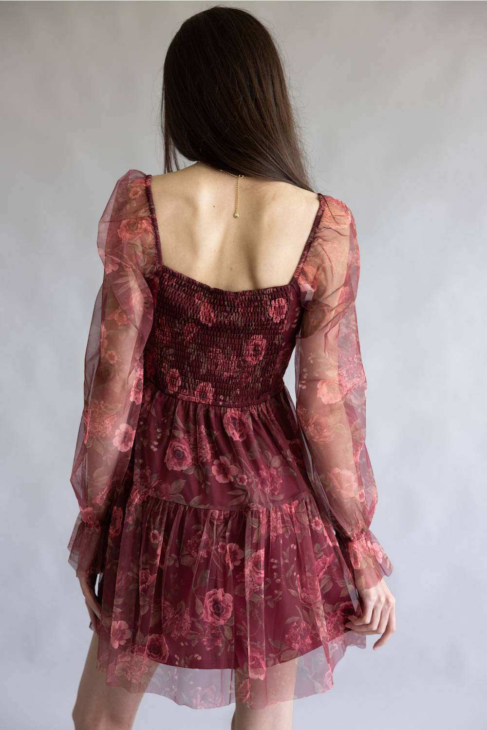 A woman viewed from behind wearing a short burgundy tulle dress with a floral pattern, sheer long sleeves, and a smocked bodice, set against a plain gray studio background.