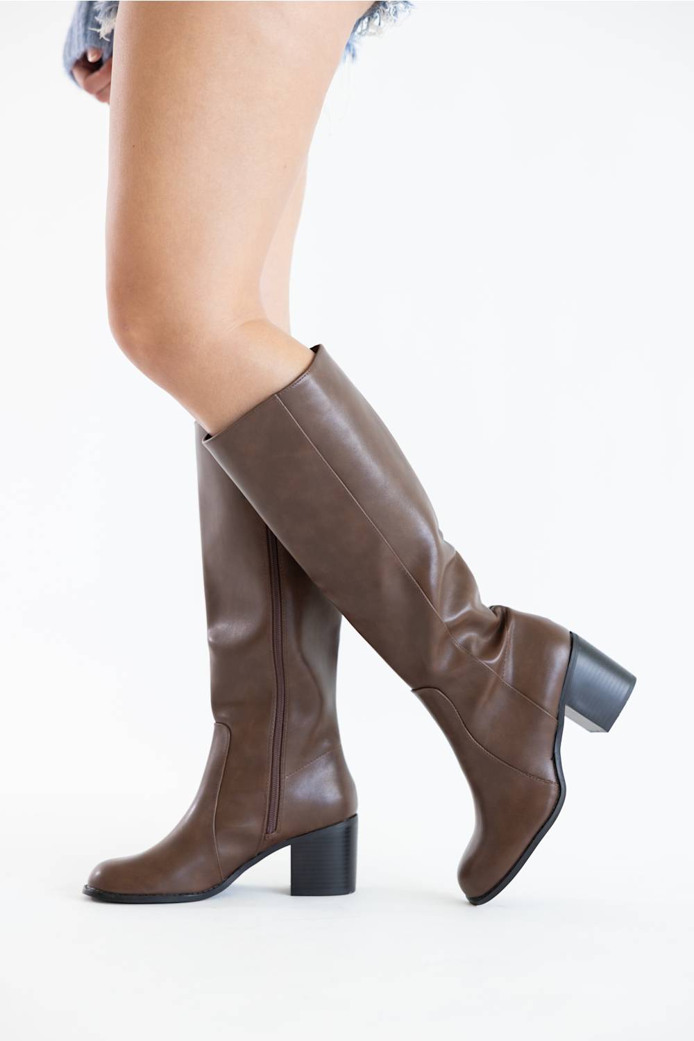 A pair of knee-high brown faux-leather boots with block heels, shown on a model's legs against a plain white studio background.