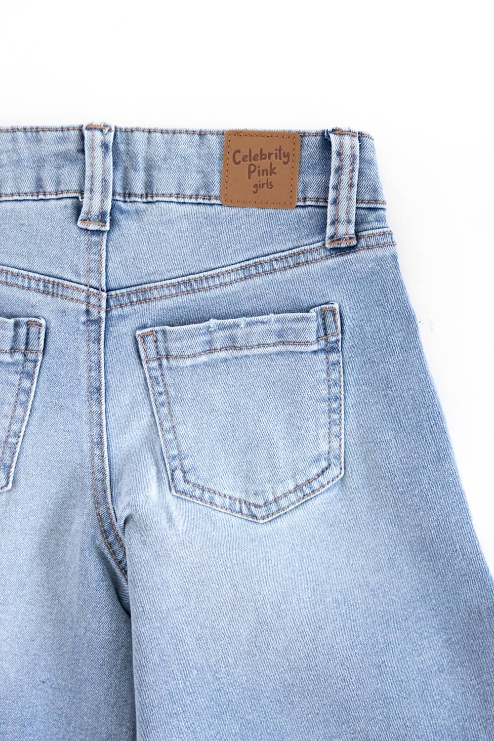 Close-up of the back waistband and right rear pocket of light-wash blue denim jeans, showing a brown leather patch with the text 'Celebrity Pink Girls' on a white background.