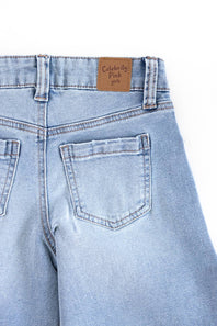 Close-up of the back waistband and right rear pocket of light-wash blue denim jeans, showing a brown leather patch with the text 'Celebrity Pink Girls' on a white background.