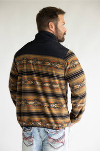 A close-up of a person wearing a quarter-zip fleece pullover with a black collar and a Southwestern geometric pattern in beige, brown, yellow, and orange. The brand name 'ARIAT' is visible on a small patch on the chest.