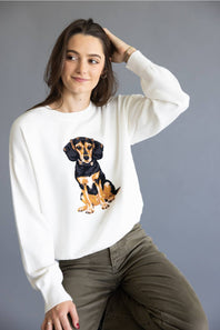 A woman wearing a white crewneck sweatshirt with a graphic of a black and tan dachshund, paired with olive green pants, posed against a solid gray studio background.