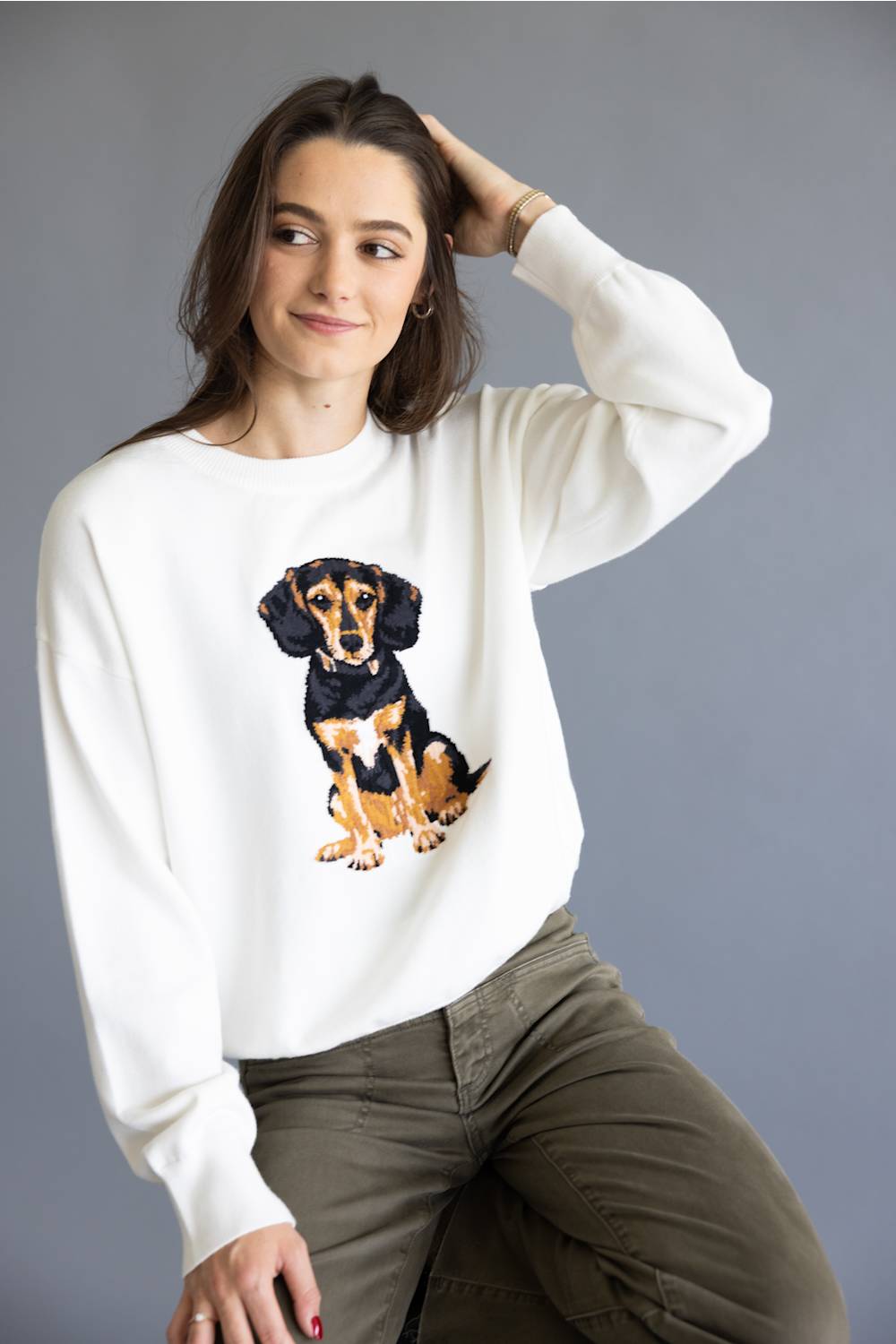 A woman wearing a white crewneck sweatshirt with a graphic of a black and tan dachshund, paired with olive green pants, posed against a solid gray studio background.