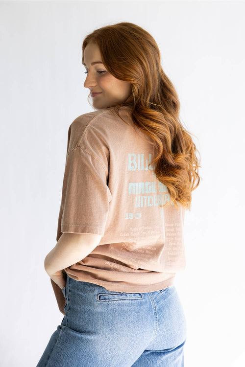 A woman with long wavy red hair is shown from the back wearing a faded light brown t-shirt with a light blue graphic print and blue jeans, set against a plain white studio background.