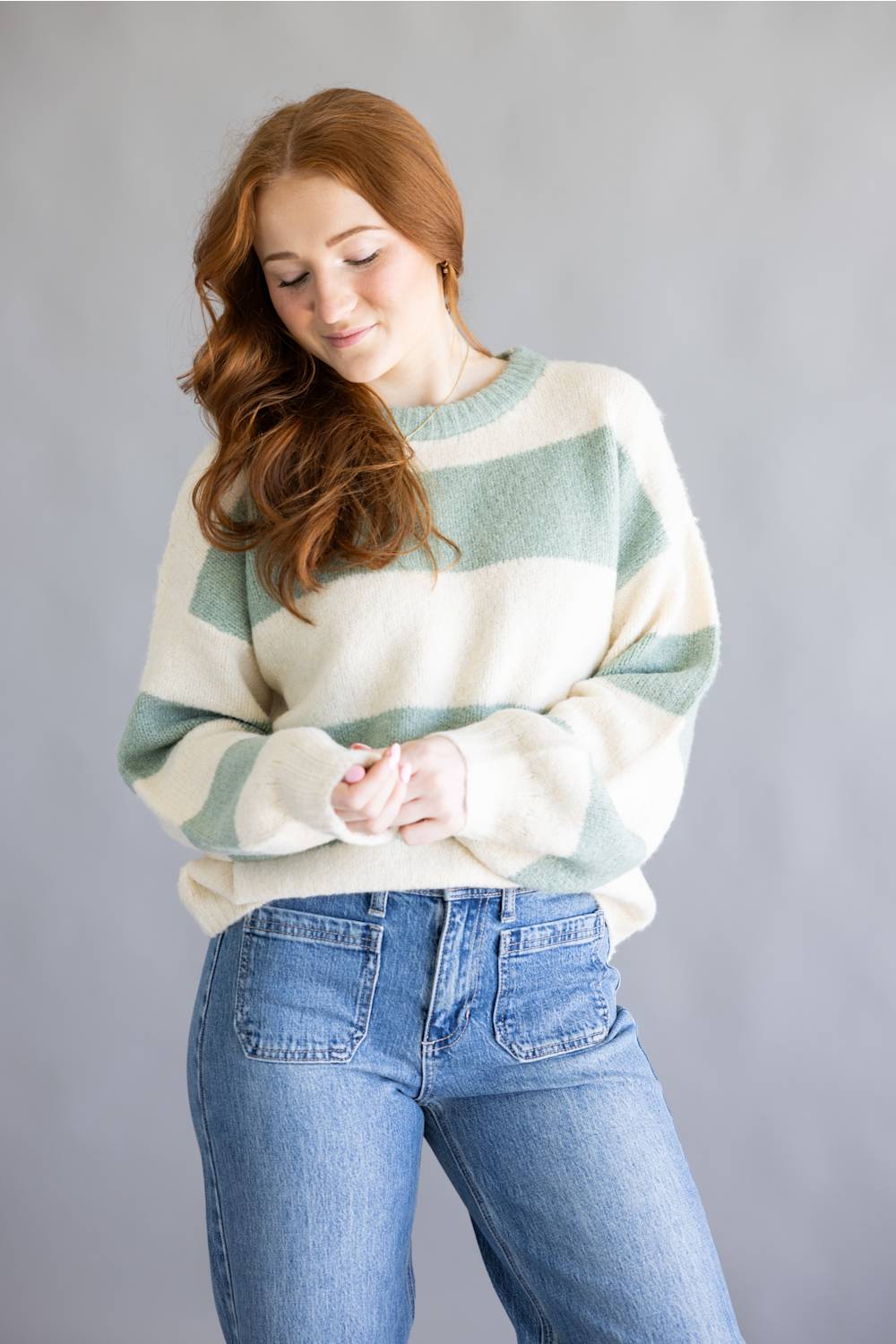 A woman with long wavy red hair wearing a fuzzy, oversized crewneck sweater with horizontal sage green and cream stripes, paired with blue jeans, standing against a solid gray studio background.