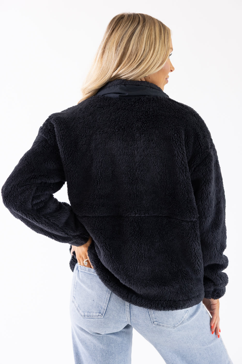 A woman shown from the back wearing a black sherpa fleece jacket and light blue jeans against a plain white background.