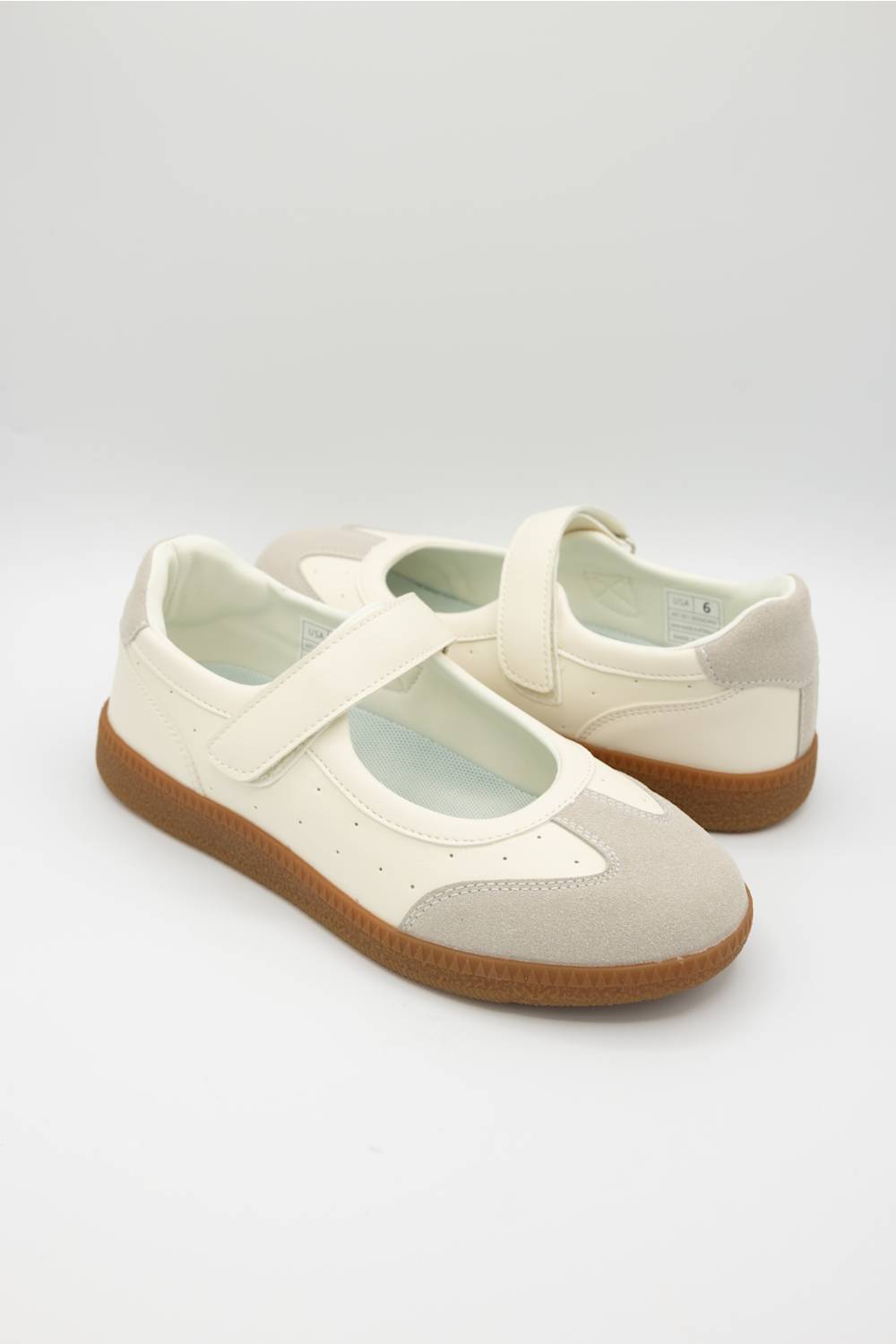 A pair of Mary Jane style shoes in off-white and beige suede, featuring a brown gum sole, photographed on a light gray studio background.