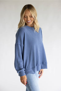 A woman wearing an oversized dusty blue crewneck sweatshirt and light wash jeans, standing against a neutral light gray studio background.