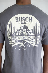 A person wearing a heather gray t-shirt, viewed from the back. The shirt features a large white circular graphic on the back with the text 'BUSCH LIGHT' above a desert landscape illustration containing saguaro cacti and mountains.