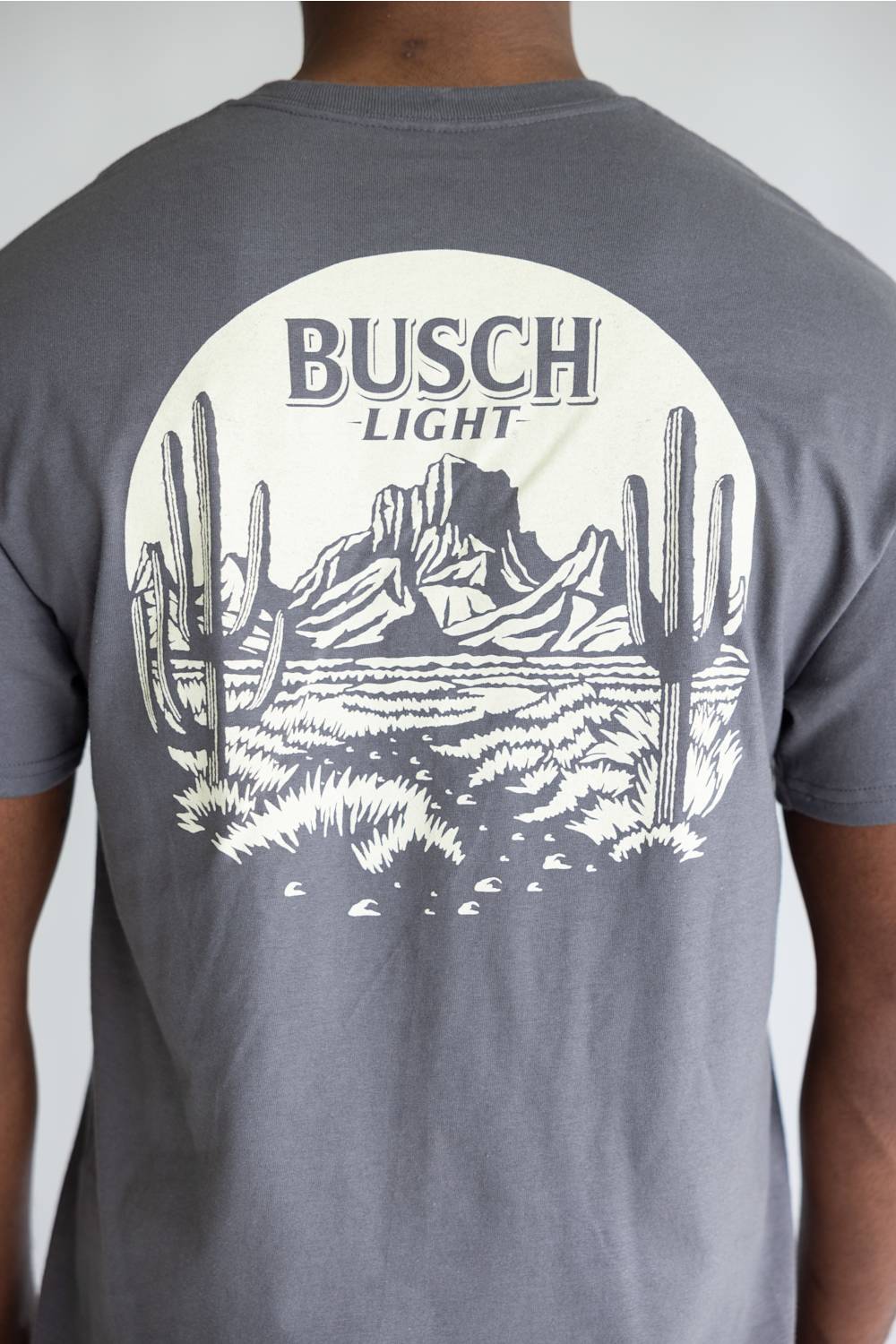 A person wearing a heather gray t-shirt, viewed from the back. The shirt features a large white circular graphic on the back with the text 'BUSCH LIGHT' above a desert landscape illustration containing saguaro cacti and mountains.