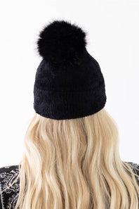 Back view of a woman with long blonde hair wearing a black plush pom-pom beanie featuring a cuffed hem, paired with a black and white patterned cardigan.
