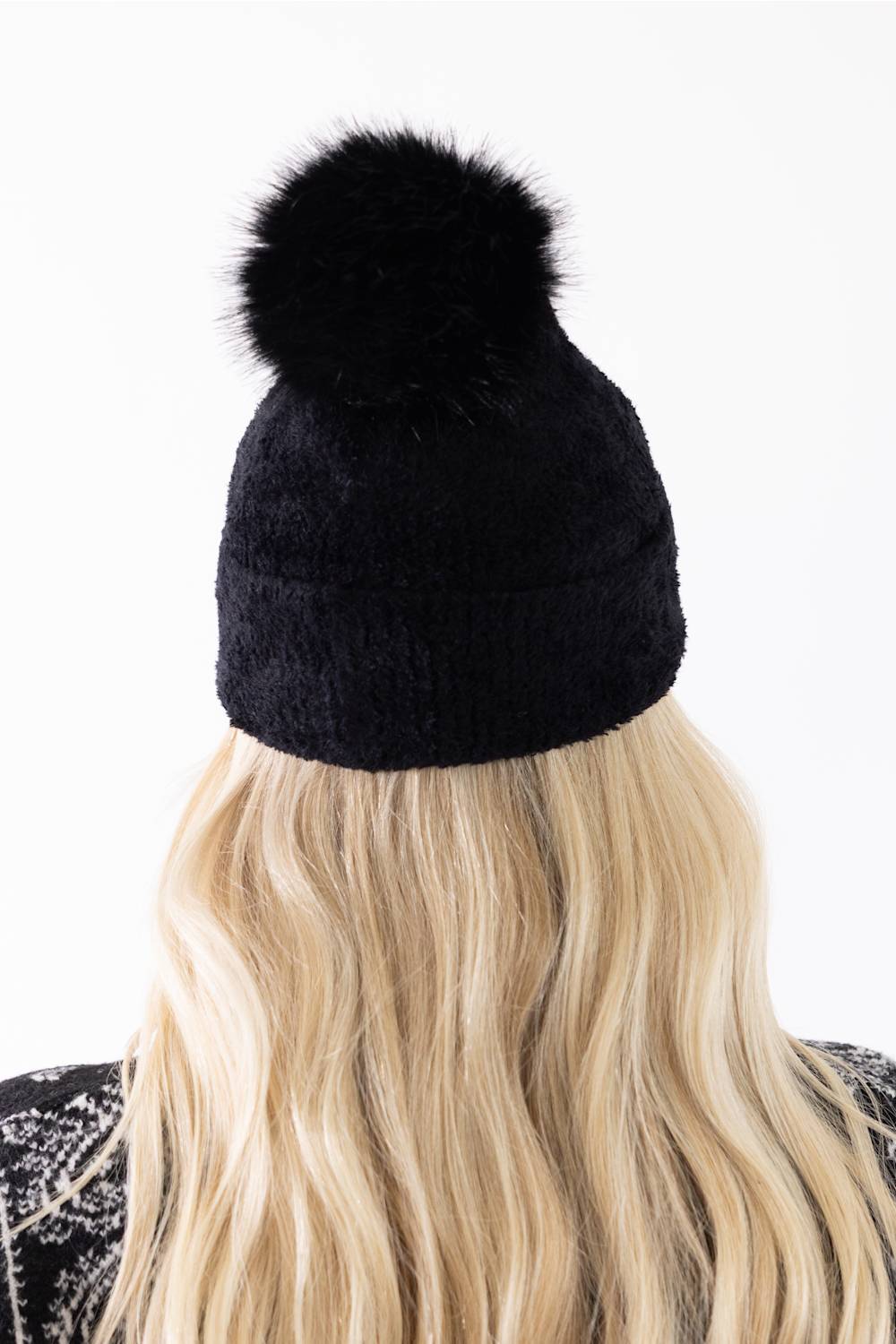 Back view of a woman with long blonde hair wearing a black plush pom-pom beanie featuring a cuffed hem, paired with a black and white patterned cardigan.