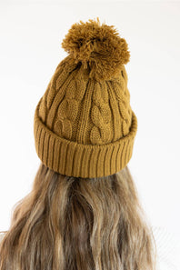 A mustard yellow cable-knit beanie with a matching pom-pom, shown on a model from the back against a plain white background.
